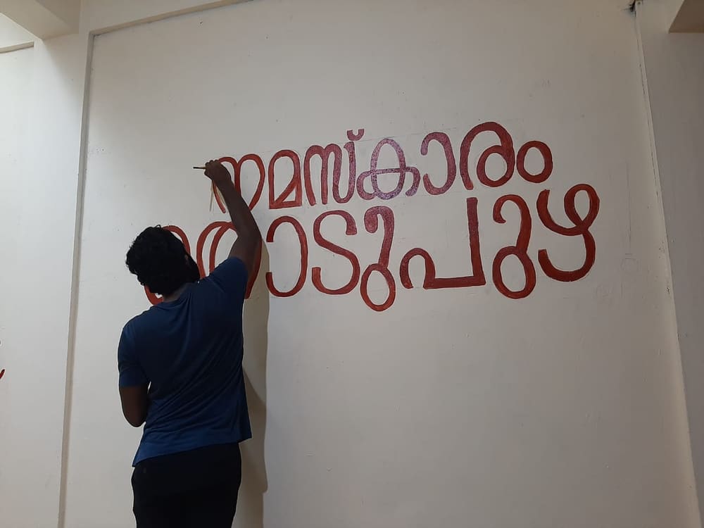 Namaskaram Thodupuzha being hand-painted in red Malayalam lettering on a white wall, the painter working on the last letters
