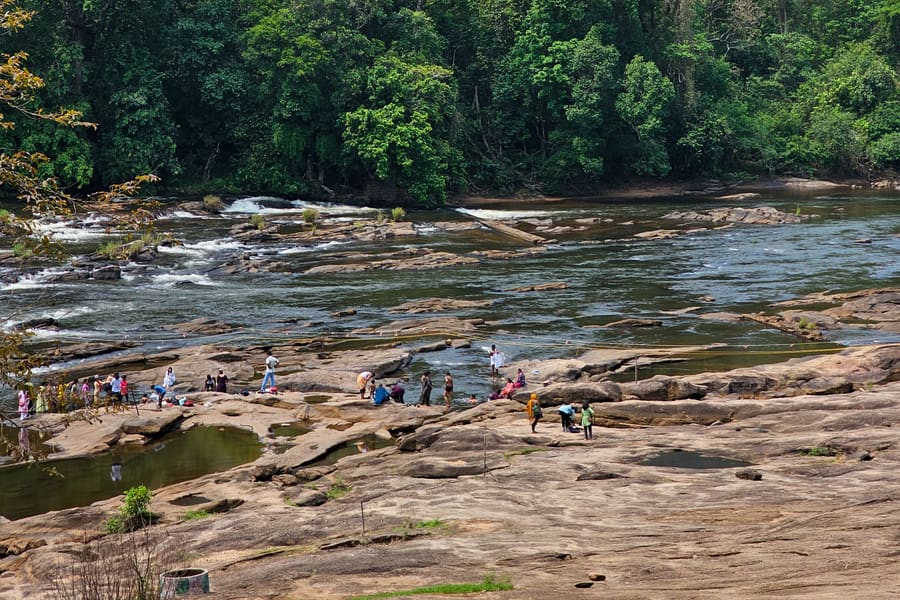 Visitors on the wide rock bed beside the stream at Thommankuthu, with forest on the far bank